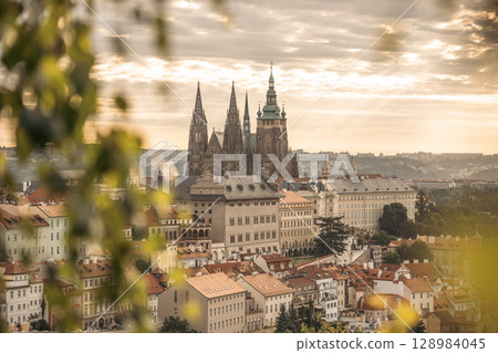 Sunlight gently illuminates Prague Castle as dawn breaks over the city, casting a warm glow on historic architecture and the sprawling rooftops of Prague. A serene atmosphere envelops the scene. Sunlight gently illuminates Prague Castle as dawn breaks over the city, casting a warm glow on historic architecture and the sprawling rooftops of Prague. A serene atmosphere envelops the scene. 128984045