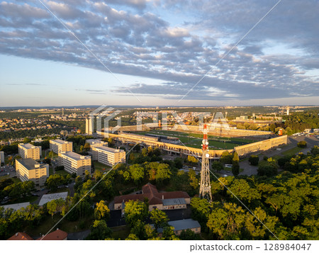 Great Strahov Stadium, surrounded by lush greenery, showcases its vastness during sunset in Prague. Buildings and trees create a beautiful landscape, highlighting the stadium's architectural grandeur. 128984047