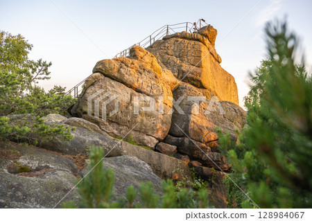 The sun sets over Popov Cliffs, casting a warm glow on the sandstone rock formations. Lush greenery surrounds the cliffs, creating a tranquil evening atmosphere in Czechia. The sun sets over Popov Cliffs, casting a warm glow on the sandstone rock formations. Lush greenery surrounds the cliffs, creating a tranquil evening atmosphere in Czechia. 128984067