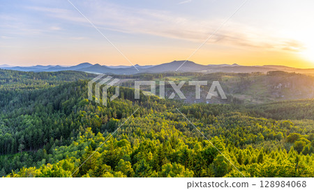 A breathtaking sunset casts golden hues over the Lusatian Mountains, viewed from the heights of Popov Cliffs. Dense forests stretch across the landscape, creating a serene evening atmosphere. 128984068