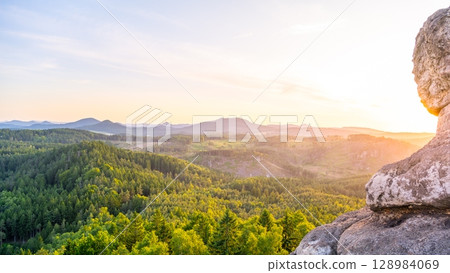 Sunlight peeks through the rocky formations at Popov Cliffs during a serene evening in the Lusatian Mountains. The sandstone cliffs are surrounded by lush greenery, creating a tranquil atmosphere. 128984069