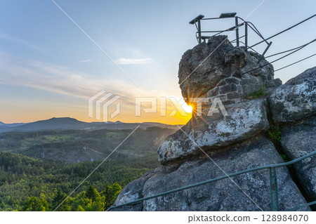Sunlight peeks through the rocky formations at Popov Cliffs during a serene evening in the Lusatian Mountains. The sandstone cliffs are surrounded by lush greenery, creating a tranquil atmosphere. Sunlight peeks through the rocky formations at Popov Cliffs during a serene evening in the Lusatian Mountains. The sandstone cliffs are surrounded by lush greenery, creating a tranquil atmosphere. 128984070