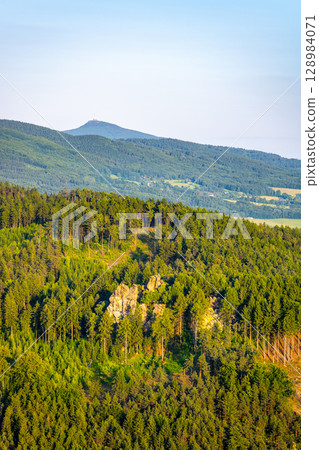 Jested Ridge is bathed in golden light as the sun sets behind the Lusatian Mountains. The sandstone cliffs rise above lush green forests, creating a breathtaking evening view. 128984071