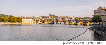 Early morning light casts a warm glow over Prague Castle and Charles Bridge. The Vltava River reflects the cityscape while the historic architecture stands majestically in the background. 128984076