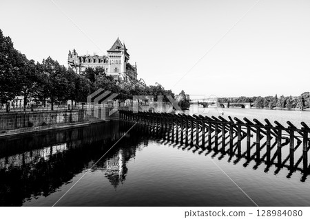Visitors stroll along Smetana Embankment in Prague, admiring the tranquil waters of the Vltava River and the historic architecture in the background on a clear day. 128984080