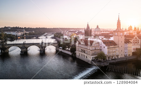 Golden light washes over Charles Bridge and the historic buildings of Prague at dawn. The serene Vltava River reflects the peaceful morning sky with no signs of activity. 128984084