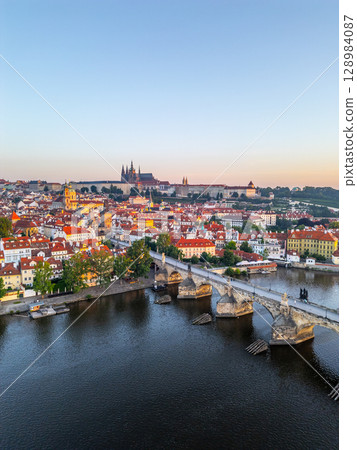 Early morning light bathes the historic city of Prague, illuminating the medieval architecture. The Charles Bridge spans the calm Vltava River, enhancing the tranquil panorama. 128984087