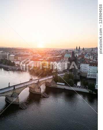 A breathtaking aerial view of Charles Bridge in Prague at sunrise. The scene showcases the Vltava River and the peaceful old town, bathed in golden morning light with no people present. 128984088