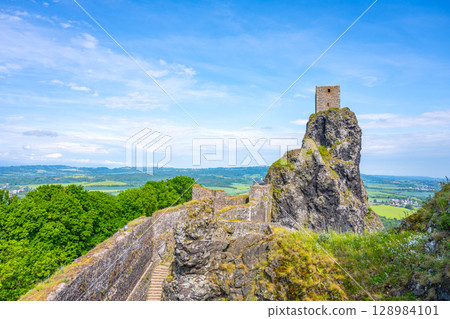 Visitors admire the historic Panna Tower perched atop a rocky outcrop at Trosky Castle Ruins in the scenic Bohemian Paradise, Czechia, surrounded by lush greenery and distant hills. 128984101