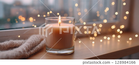 Cozy candle flame glowing in glass on wooden surface with soft knitted fabric and warm bokeh lights creating peaceful atmosphere 128984166