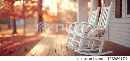 Two white rocking chairs with floral cushions sit on wooden porch bathed in warm autumn light, creating peaceful and cozy atmosphere 128984174