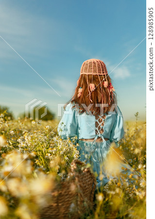 Back view girl in blue linen dress and fashionable pink crochet beanie sits in daisy field in summer day Back view girl in blue linen dress and fashionable pink crochet beanie sits in daisy field in summer day 128985905