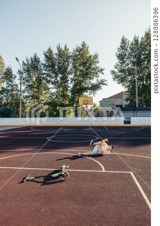 boy lying on ground after fall from scooter on wide empty basketball court with visible copy space, concept of childhood accident, outdoor play, injury awareness 128986396