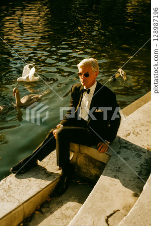 A man in a suit is sitting on a stone ledge next to a body of water 128987196