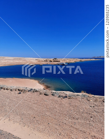 Serene Coastal Lagoon with Clear Blue Waters and Boats Under a Vibrant Blue Sky Serene Coastal Lagoon with Clear Blue Waters and Boats Under a Vibrant Blue Sky 128988235