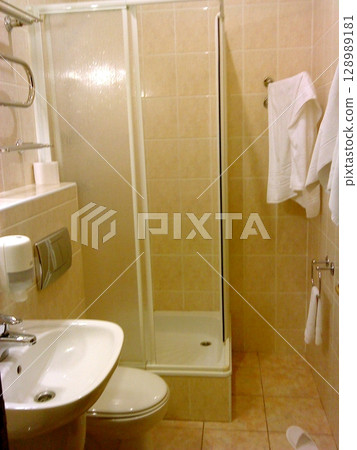A compact bathroom with beige tiles, a glass shower, white towels, a sink, and toilet, featuring a heated towel rack and slippers A compact bathroom with beige tiles, a glass shower, white towels, a sink, and toilet, featuring a heated towel rack and slippers 128989181