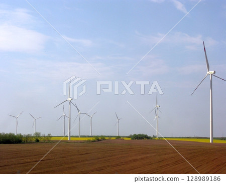 A serene landscape featuring a row of tall wind turbines stretching across a plowed field under a vast, partly cloudy sky 128989186