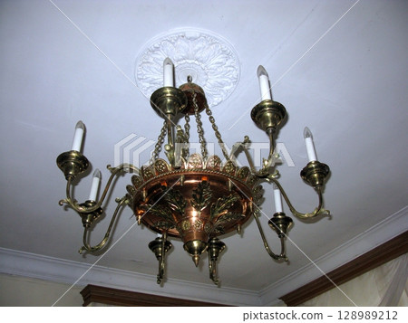 An elegant brass chandelier with candle like lights, suspended from a decorative ceiling medallion in a softly lit room with classic decor An elegant brass chandelier with candle like lights, suspended from a decorative ceiling medallion in a softly lit room with classic decor 128989212
