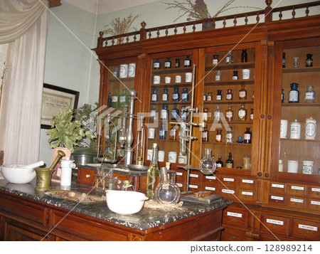 A vintage apothecary cabinet filled with glass bottles and jars, paired with a wooden counter displaying antique pharmaceutical tools A vintage apothecary cabinet filled with glass bottles and jars, paired with a wooden counter displaying antique pharmaceutical tools 128989214