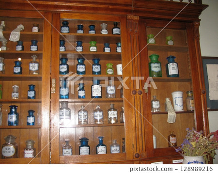 A wooden apothecary cabinet with glass doors, filled with colorful bottles and jars, accented by dried flowers in a historical setting A wooden apothecary cabinet with glass doors, filled with colorful bottles and jars, accented by dried flowers in a historical setting 128989216