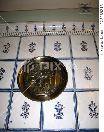 A blue and white tiled wall with a floral pattern, showcasing a brass plaque with a carved scene in a historical interior space 128989218