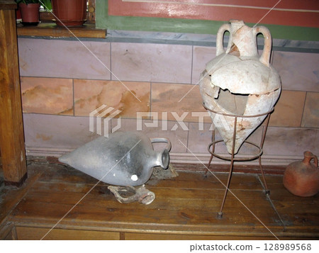 A rustic wooden surface displaying weathered ancient vases, including a cracked terracotta amphora on a stand and a darkened jug 128989568