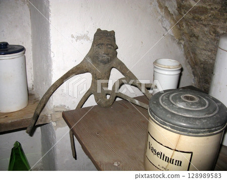 A wooden shelf displaying a carved mandrake root figure with elongated limbs, alongside ceramic jars in a stone walled historical setting A wooden shelf displaying a carved mandrake root figure with elongated limbs, alongside ceramic jars in a stone walled historical setting 128989583