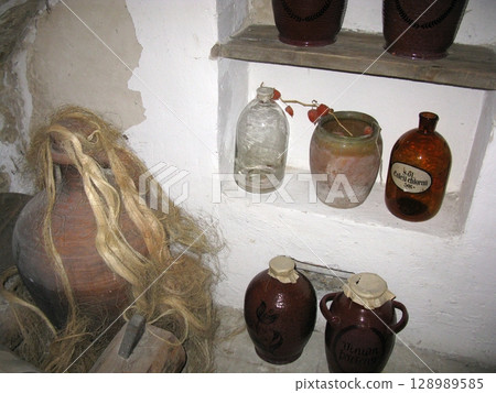 A stone walled room with a wooden shelf holding various ceramic jars, a large jug with fibers, and glass bottles in a historical display 128989585