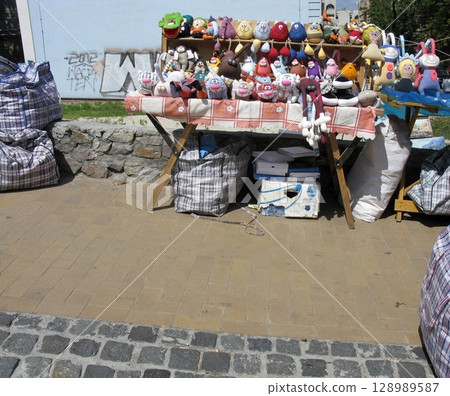 A lively outdoor market stall with a red checkered tablecloth, displaying colorful stuffed toys and various bags on a cobblestone ground A lively outdoor market stall with a red checkered tablecloth, displaying colorful stuffed toys and various bags on a cobblestone ground 128989587