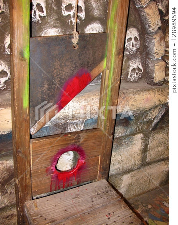 A wooden guillotine with a red stained blade, set against a stone wall embedded with skulls, creating a grim historical execution scene 128989594