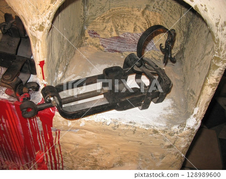 A black metal torture clamp with chains, displayed in a stone niche with red stains, evoking a dark historical punishment scene 128989600