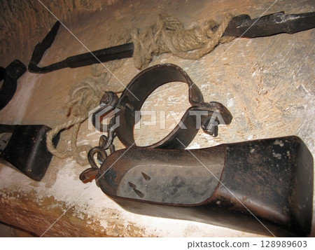 A pair of blackened metal leg screws with red stains, mounted on a wooden base in a stone walled historical torture chamber display 128989603