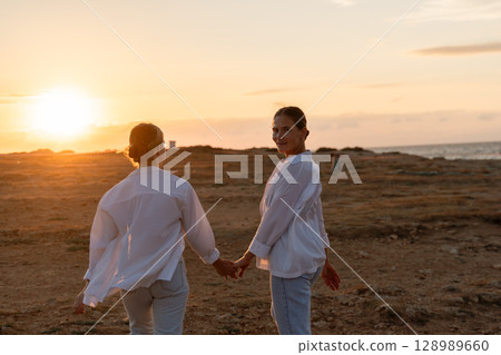 Couple Sunset Beach Walk - Two people in white clothing walk hand-in-hand towards a sunset on a beach. 128989660