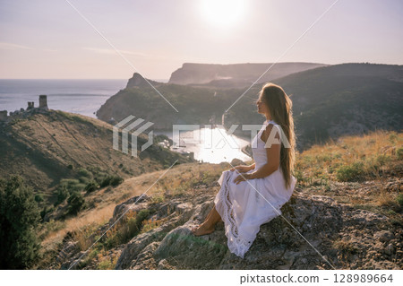 A woman in a white dress sits on a rock overlooking a body of water. The scene is serene and peaceful, with the woman enjoying the view and the calmness of the surroundings. 128989664