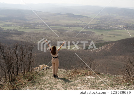 Mountains Woman Freedom: Woman stands, arms raised, overlooks valley from peak on sunny day for inspiration. 128989689
