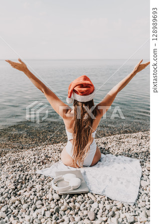 A woman wearing a red Santa hat is sitting on a towel on a beach. She is holding her arms up in the air, as if she is celebrating something. 128989693
