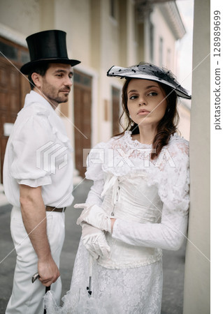 Elegant woman in white lace dress leans on column beside man in vintage attire 128989699