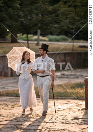Man and woman in white vintage clothes walking with lace umbrella in park 128989715