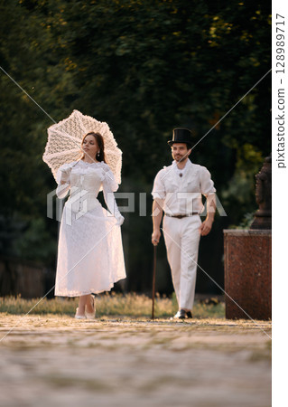 Woman walking ahead with parasol while man in top hat follows behind 128989717