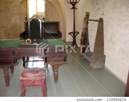 A historical office with a green topped wooden desk, a throne like chair, and a rifle rack against a brick wall with a small window A historical office with a green topped wooden desk, a throne like chair, and a rifle rack against a brick wall with a small window 128989829