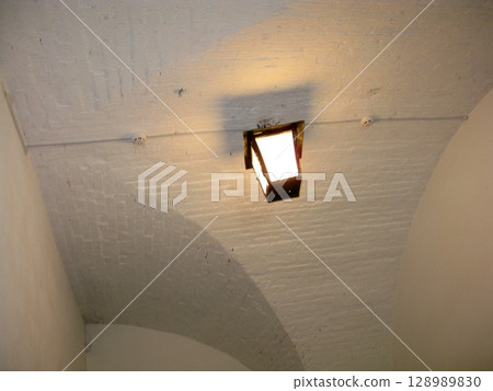 A rustic lantern hanging from a curved brick ceiling, casting a soft glow and subtle shadows in a historical interior space 128989830