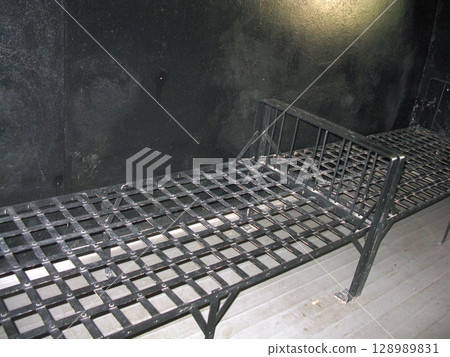 A dimly lit prison cell with two metal grid beds against a black wall, creating a somber historical incarceration scene with minimal light 128989831
