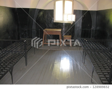 A stark prison cell with two iron framed beds, a wooden desk, and a stool under a small window, set in a historical confinement space 128989832