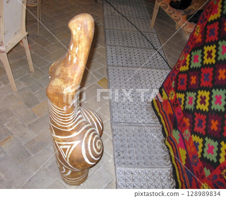 A beautifully crafted wooden sculpture with intricate white geometric patterns stands prominently on a tiled floor, complemented by a vibrant, colorful textile nearby A beautifully crafted wooden sculpture with intricate white geometric patterns stands prominently on a tiled floor, complemented by a vibrant, colorful textile nearby 128989834