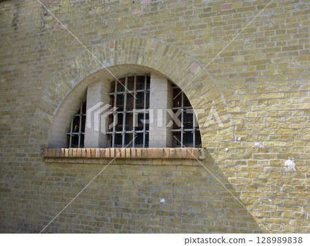 An arched brick window with metal bars on a weathered brick wall, casting shadows and highlighting historical architectural details 128989838