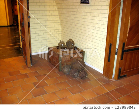 A historical exhibit of cannonballs on a wooden chest, with additional cannonballs on the floor, set against a brick wall and wooden door 128989842