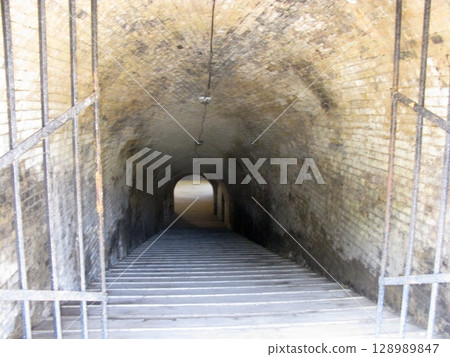 A narrow brick tunnel with a steep staircase descending into darkness, enclosed by metal railings in a historical underground setting 128989847