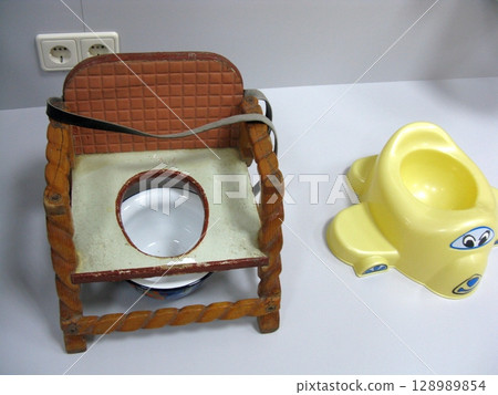 A vintage wooden child's chamber pot with a padded backrest, paired with a yellow plastic potty featuring a cartoonish face on a white surface A vintage wooden child's chamber pot with a padded backrest, paired with a yellow plastic potty featuring a cartoonish face on a white surface 128989854