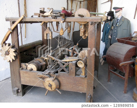 A rustic wooden hand operated textile loom with intricate gears and threads, displayed alongside historical mannequins in a traditional setting A rustic wooden hand operated textile loom with intricate gears and threads, displayed alongside historical mannequins in a traditional setting 128989858