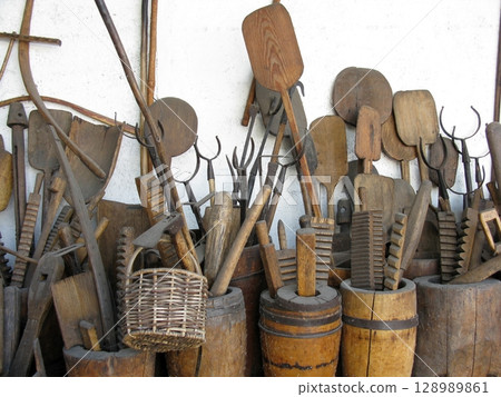 A collection of weathered wooden tools and implements arranged against a white wall with rustic barrels and a woven basket A collection of weathered wooden tools and implements arranged against a white wall with rustic barrels and a woven basket 128989861